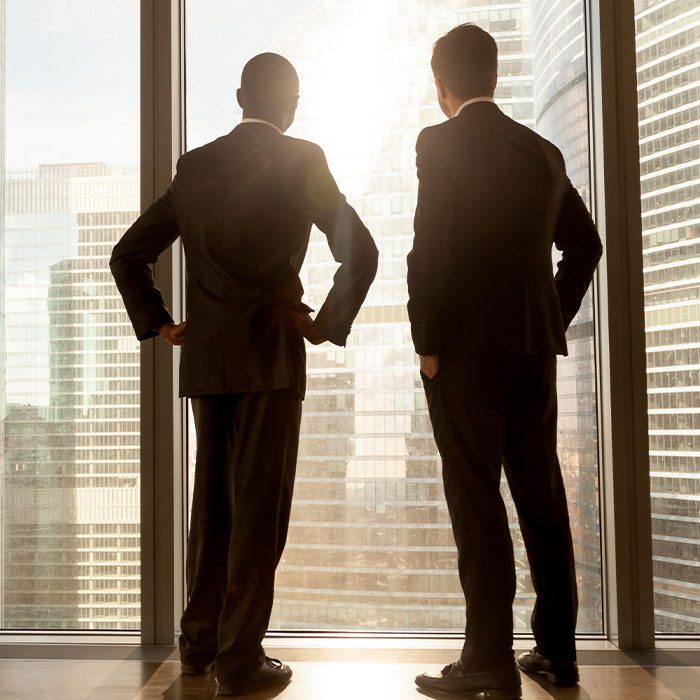 Rear view of african and caucasian businessmen wearing suits standing at full length large window enjoying great big city scenery, contemplative thoughtful multi ethnic partners building future plans