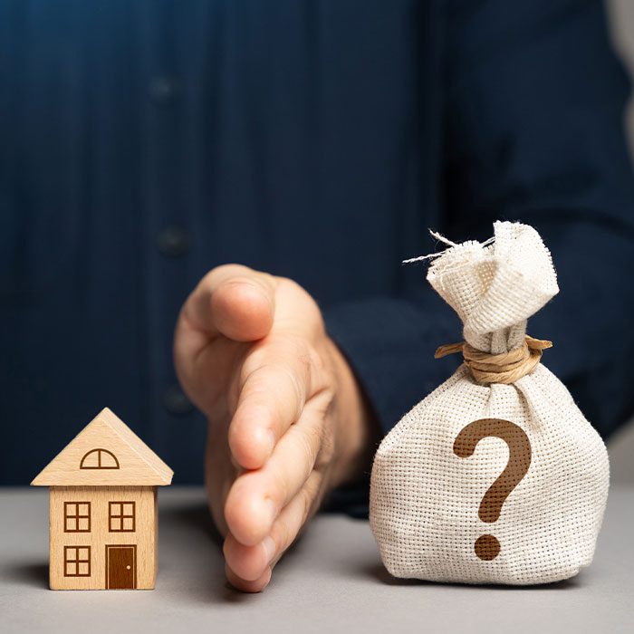 Hand in between house model and a bag of money with question mark printed on it
