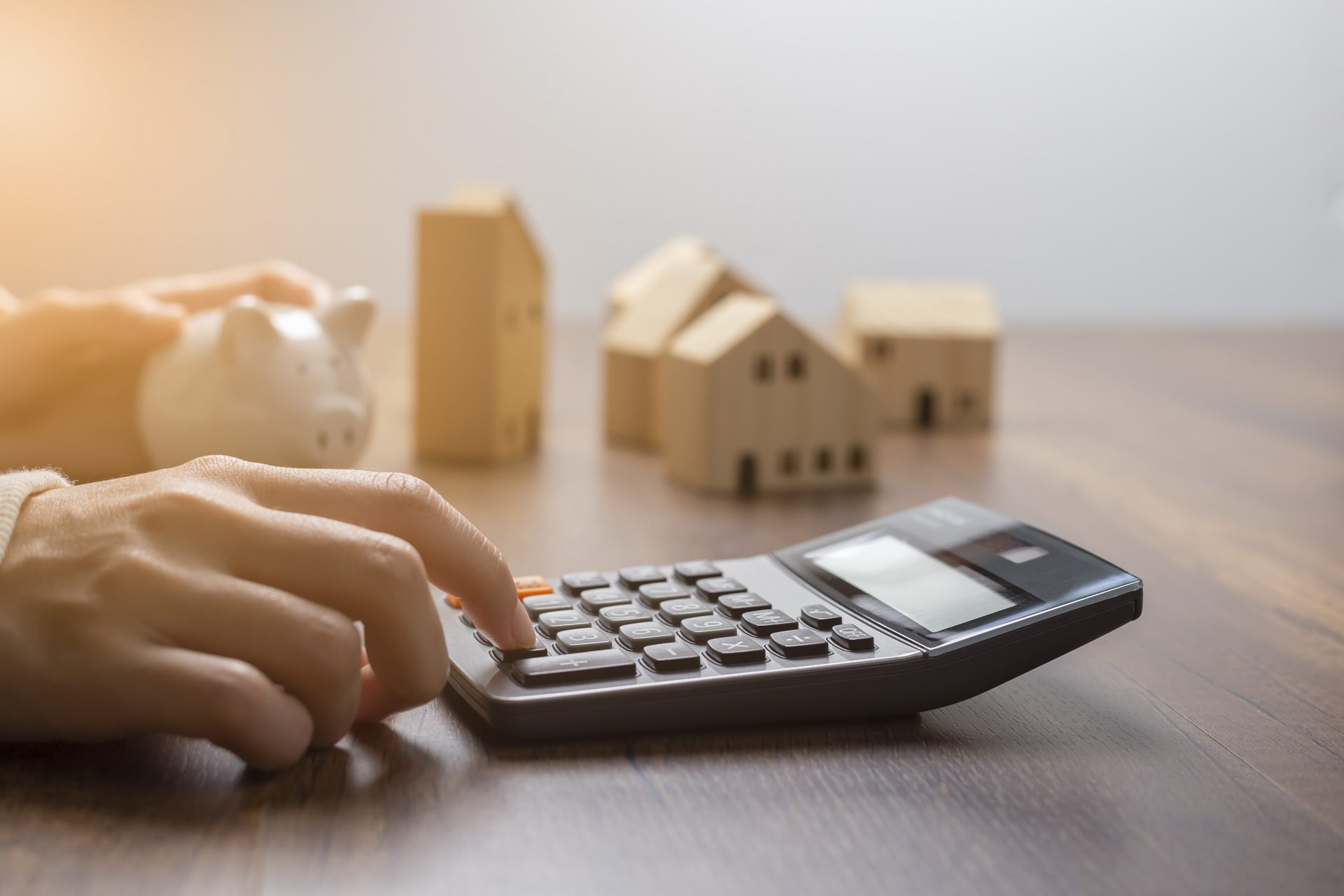 The hand is pressing calculators, piggy bank with wooden house. buy or rent question on note with calculators on desk. Property investment calculations